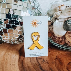 May include: Yellow enamel pin with a yellow ribbon and a white Mickey Mouse head in the center. The pin is on a white card with the text "xll wishes" and "Enamel Pin" printed on it.