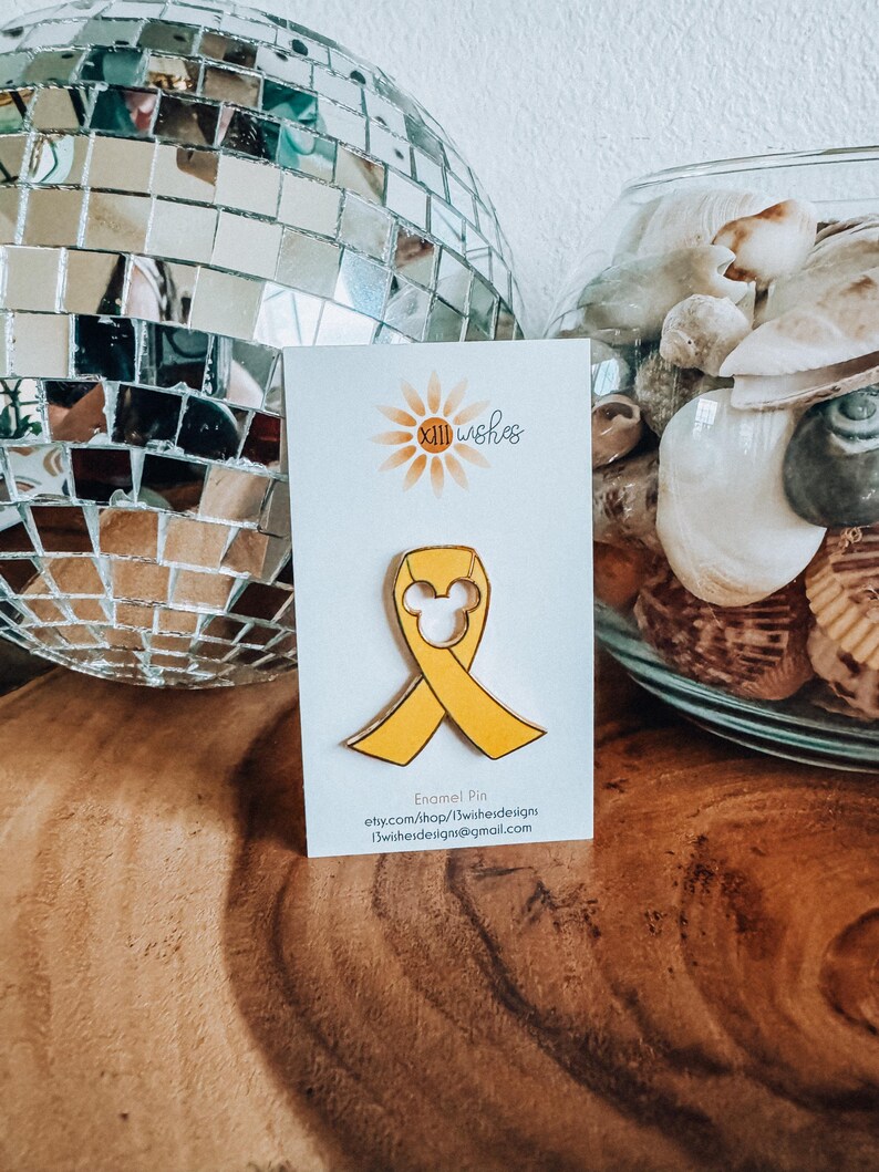 May include: Yellow enamel pin with a yellow ribbon and a white Mickey Mouse head in the center. The pin is on a white card with the text "xll wishes" and "Enamel Pin" printed on it.