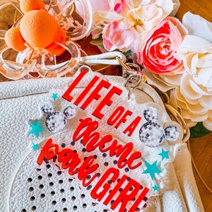 Puede incluir: Bolso blanco con dise&ntilde;o perforado, con un charm acr&iacute;lico rojo que dice "LIFE OF A Theme Park GIRL". El charm est&aacute; adornado con formas de Mickey Mouse de bola de discoteca y estrellas turquesas. La imagen tambi&eacute;n incluye pinzas para el pelo naranjas y flores coloridas.