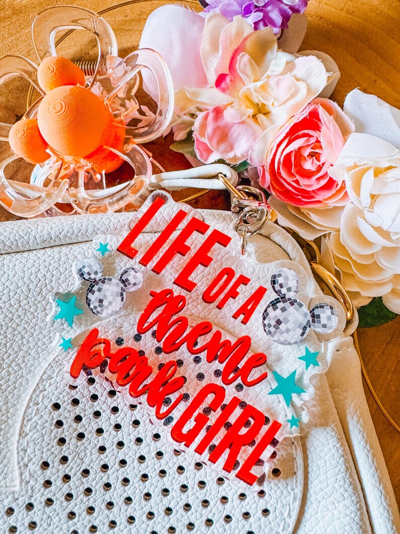 Puede incluir: Bolso blanco con dise&ntilde;o perforado, con un charm acr&iacute;lico rojo que dice "LIFE OF A Theme Park GIRL". El charm est&aacute; adornado con formas de Mickey Mouse de bola de discoteca y estrellas turquesas. La imagen tambi&eacute;n incluye pinzas para el pelo naranjas y flores coloridas.
