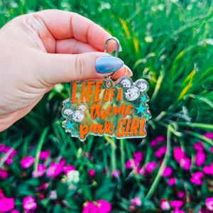 Puede incluir: Un llavero acr&iacute;lico transparente con el texto "LIFE OF A theme park GIRL" en naranja, con siluetas de cabezas de Mickey Mouse y estrellas turquesas. El llavero se sostiene frente a un fondo de follaje verde y flores rosas.