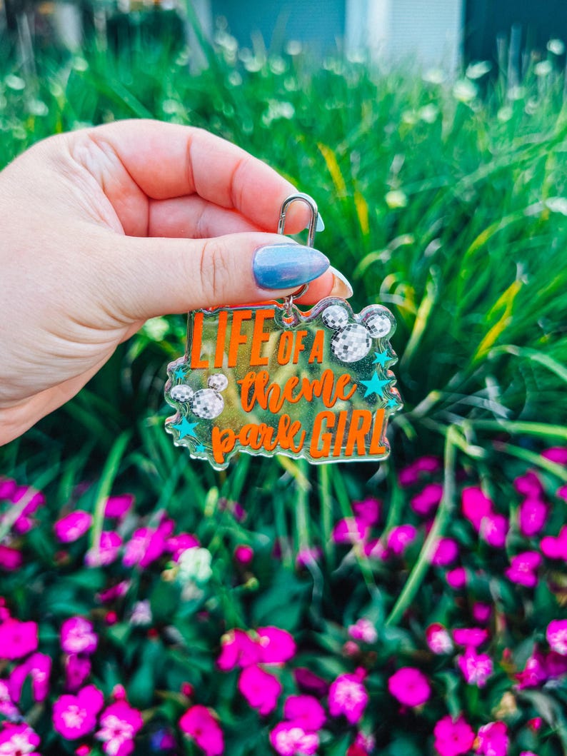 Puede incluir: Un llavero acr&iacute;lico transparente con el texto "LIFE OF A theme park GIRL" en naranja, con siluetas de cabezas de Mickey Mouse y estrellas turquesas. El llavero se sostiene frente a un fondo de follaje verde y flores rosas.