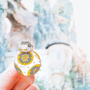 May include: A white and yellow enamel pin of a Star Wars BB-8 droid with a yellow flower design.