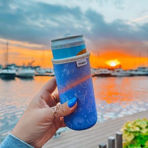 May include: A blue can cooler with a white and tan rim, featuring a pattern of seashells, cacti, and a bull skull. The cooler is holding a can with the text "FREE NO ADDED SUGAR". The background shows a sunset over a marina.