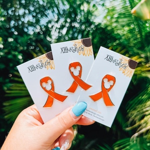 May include: Three enamel pins featuring an orange ribbon design with a Mickey Mouse head silhouette. Each pin is mounted on a white card with the text "XIII wishes" and a sunflower illustration. The pins are held against a blurred green background.
