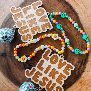 May include: Two wooden pendants with the text "LOVE LIKE JESUS" and a small disco ball attached. Colorful beaded necklaces with smiley faces, hearts, and flower beads are also present. The items are on a wooden surface.
