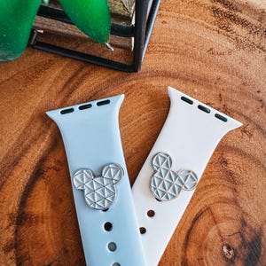 May include: Two silicone watch straps, one light blue and one white, each featuring a silver Mickey Mouse-shaped charm. The charms have a geometric design. The straps are displayed on a wooden surface.