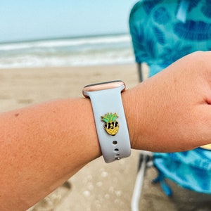 May include: A light blue silicone Apple Watch band with a gold pineapple pin with sunglasses. The watch is on a person's wrist at the beach.