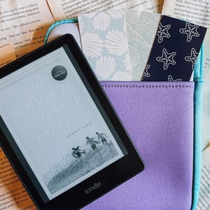 May include: A Kindle e-reader showing "The Summer I Turned Pretty" by Jenny Han, on a purple and turquoise case. The case contains patterned bookmarks with seashell and starfish designs. The scene is set on a background of open books.