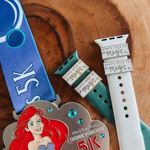 May include: A white and teal blue running medal with a picture of Ariel from The Little Mermaid. The medal says "Disney Princess 5K" and has a blue ribbon attached.  Two gold watch bands with the text "every mile is magic" are also in the image.