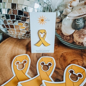 May include: Yellow enamel pin featuring a Mickey Mouse head in the center of a yellow ribbon. The pin is on a white card with the text "XIII Wishes" and a small sunflower graphic. The text "Enamel Pin" is also on the card, along with the shop's website and email address.