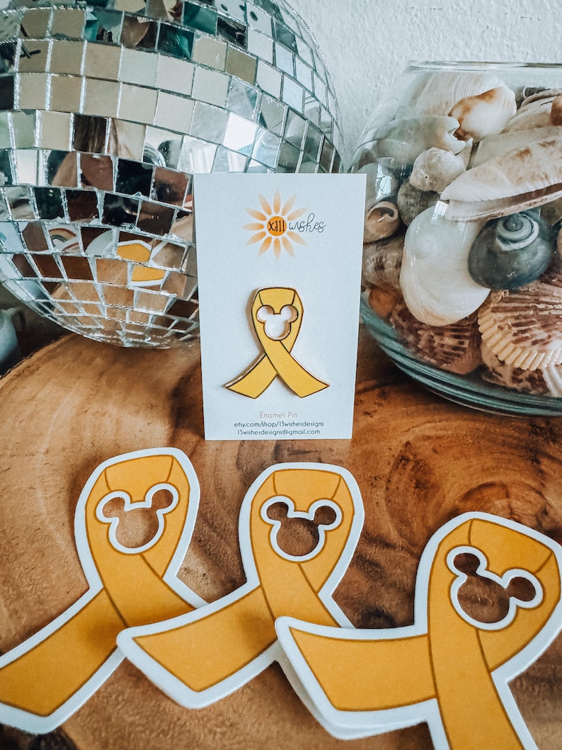 May include: Yellow enamel pin featuring a Mickey Mouse head in the center of a yellow ribbon. The pin is on a white card with the text "XIII Wishes" and a small sunflower graphic. The text "Enamel Pin" is also on the card, along with the shop's website and email address.