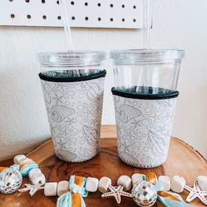 May include: Two clear plastic cups with black and white sea-themed sleeves. The sleeves feature a pattern of seashells, starfish, and bubbles. The cups are on a wooden surface with a blue and orange tassel garland.