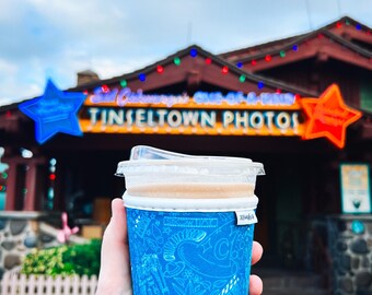 May include: A blue coffee cup sleeve with a tropical pattern, held in front of a building with the text "TINSELTOWN PHOTO". The cup contains a beverage and has a clear plastic lid. The building has star-shaped neon signs.