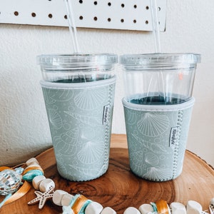 May include: Two clear plastic tumblers with lids and straws. The tumblers are in light blue insulated sleeves with a white seashell pattern. A decorative garland with wooden beads, starfish, and a disco ball is in the foreground.