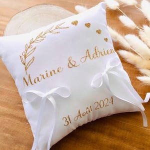May include: White pillow with gold glitter embroidery. The pillow has a floral design, the names "Marine & Adrien" and the date "31 Août 2024".