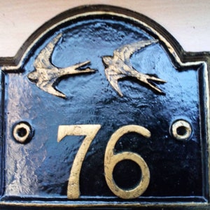 Puede incluir: Placa de número de casa de metal negro con el número 76 pintado en oro y dos pájaros dorados en vuelo.