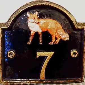Könnte beinhalten: Ein schwarzes und goldenes Hausnummernschild mit einer Fuchs-Illustration. Die Nummer 7 ist in Gold auf schwarzem Hintergrund. Der Fuchs ist orange und weiß und blickt nach rechts.