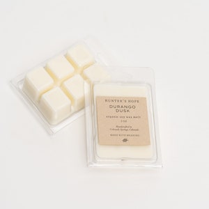Op de afbeelding: Een verpakking van doorzichtig plastic met zes witte sojawas smelttabletten. Het etiket vermeldt "Hunter's Hope Durango Dusk organic soy wax melt 3 oz Handcrafted in Colorado Springs, Colorado MADE WITH MEANING".