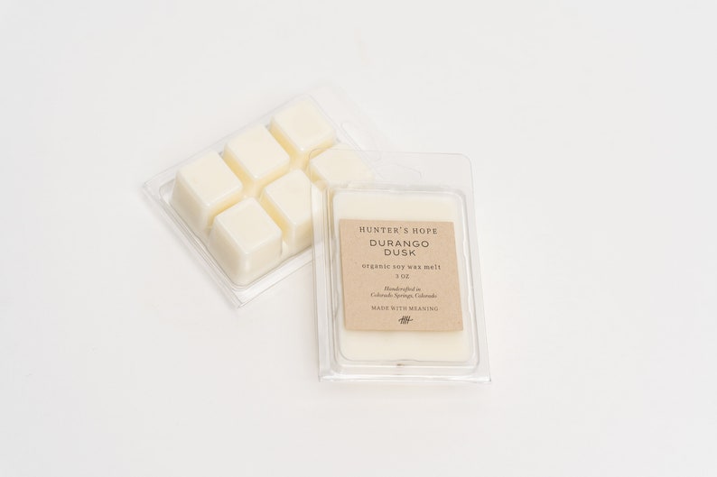Op de afbeelding: Een verpakking van doorzichtig plastic met zes witte sojawas smelttabletten. Het etiket vermeldt "Hunter's Hope Durango Dusk organic soy wax melt 3 oz Handcrafted in Colorado Springs, Colorado MADE WITH MEANING".