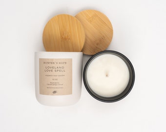 Op de afbeelding: Een witte kaars met een zwarte deksel en een bruin label met de tekst "HUNTER'S HOPE LOVELAND LOVE SPELL biologische sojawas kaars 10 OZ Handgemaakt in Colorado Springs, Colorado MADE WITH MEANING #HH". De kaars staat op een wit oppervlak met twee houten deksels.