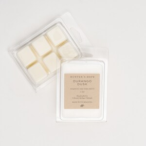 Op de afbeelding: Een doorzichtige plastic container met zes sojawas smelttabletten, gelabeld met "Hunter's Hope Durango Dusk organic soy wax melt 3 oz.  Manufactured in  Caledonia, Springs, Colorado  Made with Meaning"