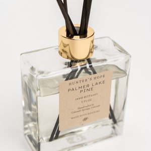 May include: A clear glass bottle with a gold lid and black reeds, filled with a clear liquid. The label reads "HUNTER'S HOPE PALMER LAKE PINE reed diffuser 5 FL OZ Handcrafted in Colorado Springs, Colorado MADE WITH MEANING".