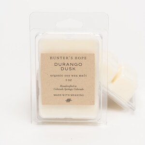 Op de afbeelding: Een kleine, witte, organische sojawas smelt in een doorzichtige plastic verpakking. Het etiket vermeldt "Hunter's Hope Durango Dusk organic soy wax melt 3 oz Handcrafted in Colorado Springs, Colorado MADE WITH MEANING 丰".