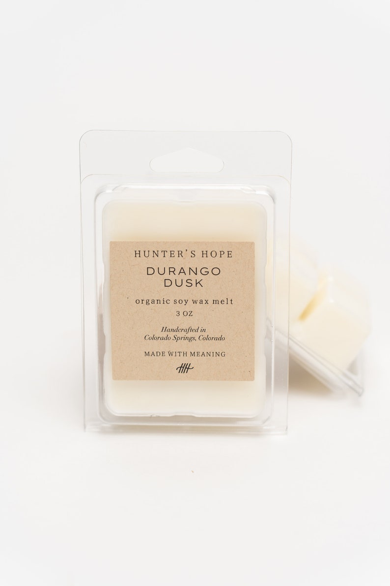 Op de afbeelding: Een kleine, witte, organische sojawas smelt in een doorzichtige plastic verpakking. Het etiket vermeldt "Hunter's Hope Durango Dusk organic soy wax melt 3 oz Handcrafted in Colorado Springs, Colorado MADE WITH MEANING 丰".