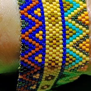 May include: A beaded bracelet with a colorful, geometric design. The bracelet features rows of small, evenly spaced beads in shades of blue, orange, yellow, and turquoise. The bracelet is approximately 1 inch wide.