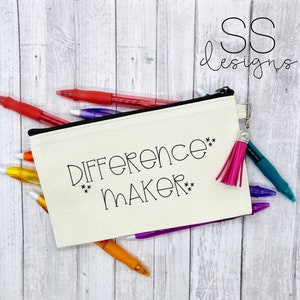 Peut inclure: Une trousse à crayons en toile blanche avec une fermeture éclair noire et un pompon rose. La trousse porte l'inscription "Difference Maker" en noir.
