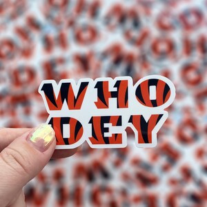 Op de afbeelding: Een sticker met de woorden "WHO DEY" in oranje en zwarte tijgerstrepen. De sticker wordt vastgehouden door een hand.