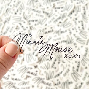 Op de afbeelding: Een stapel transparante stickers met de tekst "Minnie Mouse XOXO" in zwarte schrift. De stickers liggen op een wit oppervlak.