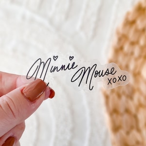 Op de afbeelding: Transparante sticker met de tekst "Minnie Mouse xoxo" in zwarte cursieve letters. Twee kleine hartjes bevinden zich boven het woord "Minnie".