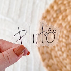 May include: A clear sticker with the word "Pluto" written in black cursive lettering. The sticker has a paw print design.