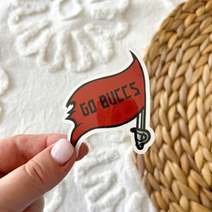Może przedstawiać: Naklejka w kolorze czerwonym i białym z motywem pirackiej flagi. Flaga ma biały miecz i napis "GO BUCCS" w kolorze czarnym.