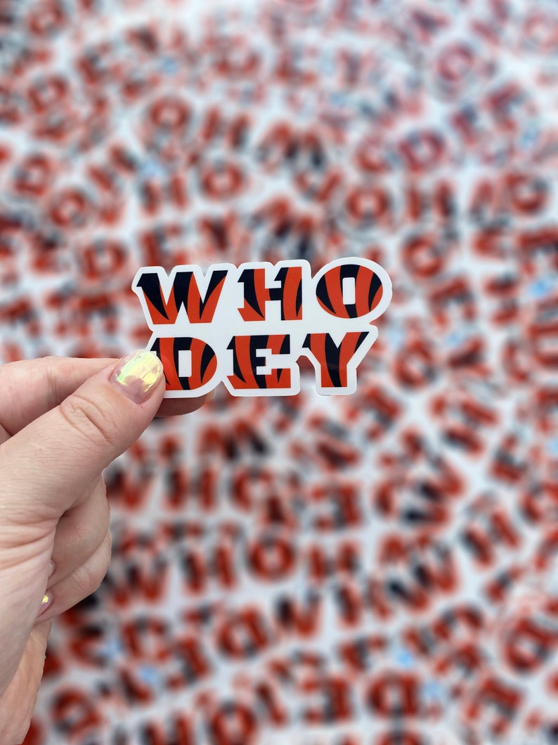 Op de afbeelding: Een sticker met de woorden "WHO DEY" in oranje en zwarte tijgerstrepen. De sticker wordt vastgehouden door een hand.