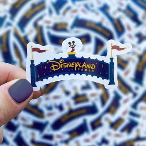 May include: A Disneyland Paris sticker. The design features a blue and gold arched sign with the Disneyland logo and the word "Paris". Mickey Mouse is on top of the sign. Two flags with red banners are on either side.