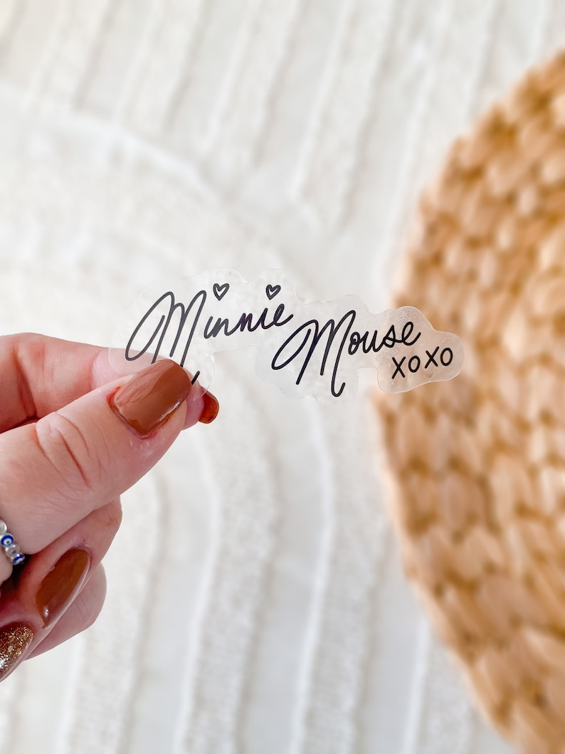 Op de afbeelding: Transparante sticker met de tekst "Minnie Mouse xoxo" in zwarte cursieve letters. Twee kleine hartjes bevinden zich boven het woord "Minnie".