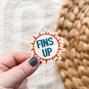 May include: A white sticker with an orange sunburst design. The sticker says "FINS UP" in teal blue letters.