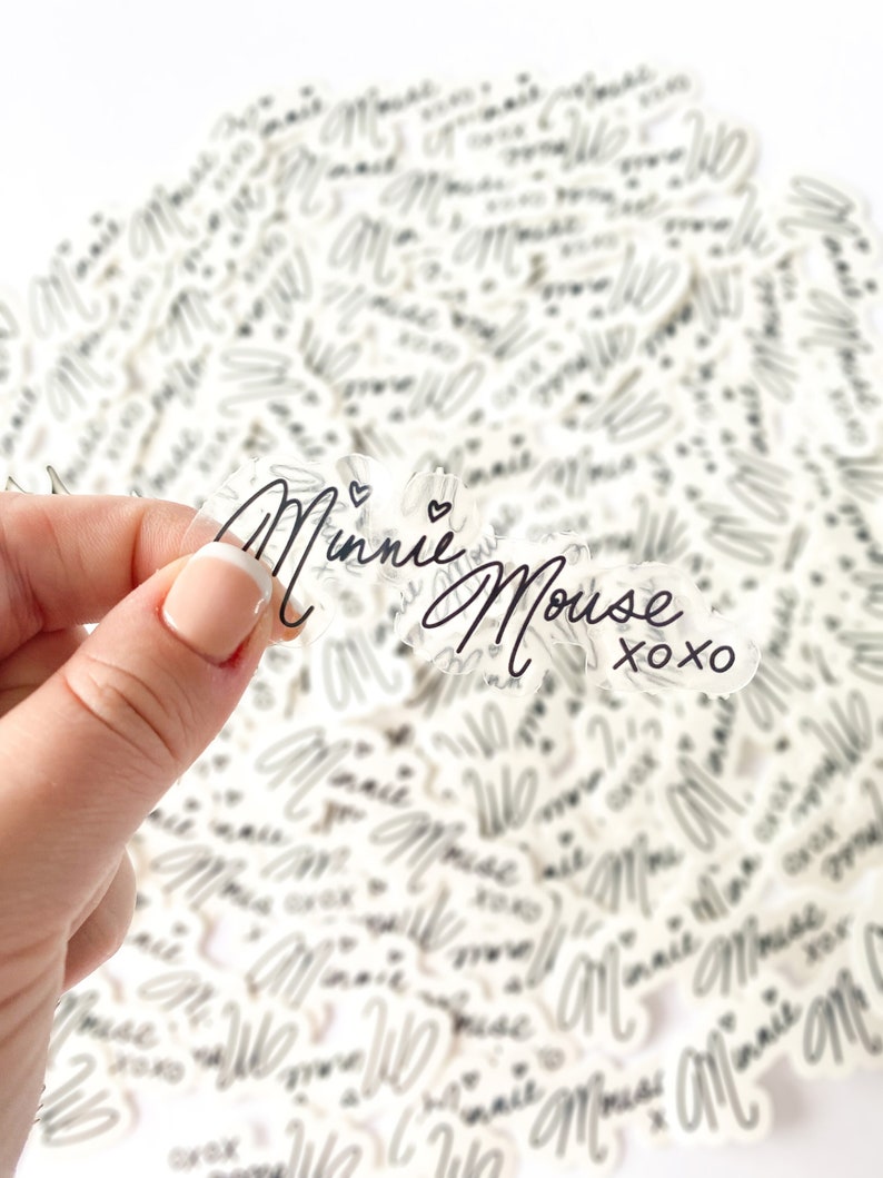 Op de afbeelding: Een stapel transparante stickers met de tekst "Minnie Mouse XOXO" in zwarte schrift. De stickers liggen op een wit oppervlak.