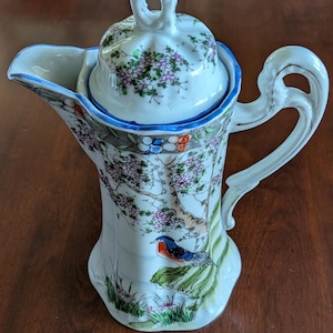 May include: White porcelain pitcher with a lid, featuring a blue rim and a handle. The pitcher is decorated with a floral and bird design in shades of pink, blue, and green. The pitcher is approximately 8 inches tall.