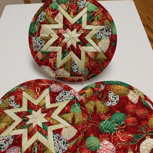 May include: Three round Christmas-themed quilted trivets. The trivets feature a red background with gold, green, and white holiday patterns. Each trivet has a central star design in cream and gold. The trivets are approximately 8 inches in diameter.