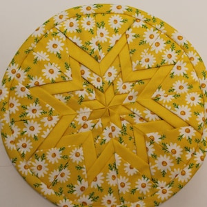 May include: A circular quilted potholder featuring a folded fabric star pattern. The design incorporates a yellow fabric with a white daisy print and solid yellow fabric accents, creating a geometric star shape radiating from the center.