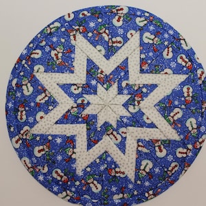 May include: A round, quilted Christmas trivet with a blue background covered in snowmen and snowflakes. A white star is appliqued in the center. The trivet is approximately 9 inches in diameter.