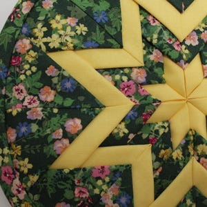 May include: A circular quilted fabric ornament featuring a folded star pattern. The design alternates between solid yellow fabric strips and a dark green floral print fabric with small pink, blue, and yellow flowers throughout the surface.