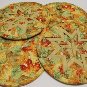 May include: Four round quilted pot holders with a fall leaf pattern in shades of orange, yellow, and green. Each pot holder features a tan starburst design in the center. Kitchen decor.