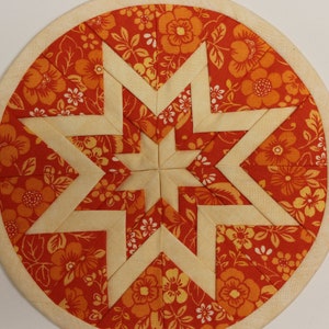 May include: A circular fabric wall hanging featuring a geometric star pattern. The design consists of cream-colored fabric strips forming a central star, surrounded by orange and red floral patterned fabric segments, all framed by a light wood hoop.