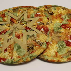 May include: Two round quilted pot holders. One features a tan starburst design, the other has a loop for hanging. Both are covered in a fall leaf pattern with shades of green, yellow, and brown.