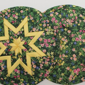 May include: Two round quilted potholders featuring a dark green fabric with a multi-colored floral print. One potholder displays a yellow eight-pointed star design in the center, while the other shows the floral pattern with a small fabric loop.